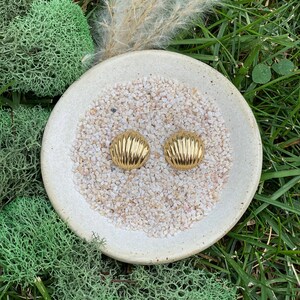 May include: A pair of gold-tone earrings with a shell-like design. The earrings are displayed on a white ceramic dish filled with small white stones.