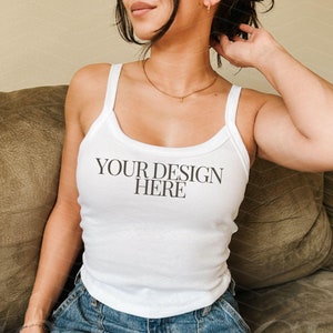 Puede incluir: Un top blanco de tirantes con el texto "YOUR DESIGN HERE" impreso en la parte delantera. El top lo lleva puesto una persona sentada en un sofá marrón.