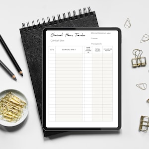 May include: A black spiral-bound notebook and a black tablet displaying a "Clinical Hours Tracker" form. Two pencils, a small dish of gold paper clips, and gold binder clips are arranged on a white surface.