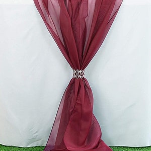 May include: Burgundy table runner with a crystal table runner holder. The runner is made of sheer fabric and is draped over a white tablecloth.