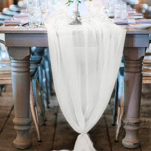 May include: White sheer table runner draped over a wooden table with white chairs. The runner is gathered at the centre and hangs down to the floor.