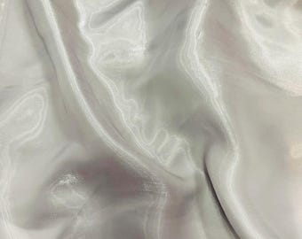 May include: A close-up of a smooth, white fabric with a subtle sheen. The fabric is draped and folded, creating soft, flowing lines.