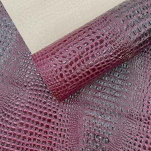 May include: A close-up of a roll of burgundy faux leather with a crocodile skin texture. The material is glossy and reflects light, with a lighter beige fabric in the background. The pattern is detailed and realistic.