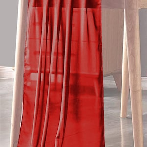 May include: A red table runner made of a soft, flowing fabric. The runner is draped over a wooden table and hangs down the sides.