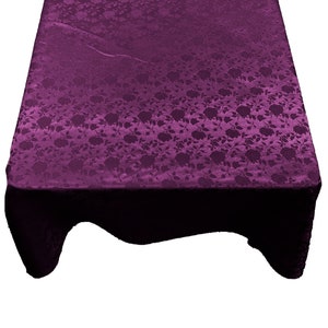 May include: A rectangular, deep purple tablecloth with a floral pattern. The fabric has a subtle sheen, and the floral design is visible across the entire surface. The tablecloth is draped over a table, with the edges hanging down.