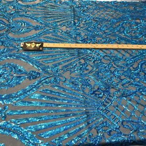 May include: A close-up of a teal sequined fabric with an intricate, ornate design. The fabric features a repeating pattern of swirling lines and floral motifs. A ruler is placed across the fabric, showing measurements in inches.