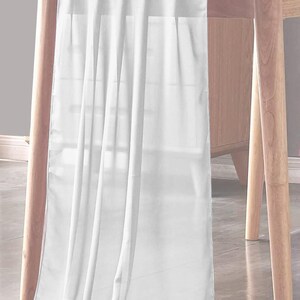 May include: White table runner made of sheer fabric, draped over a wooden table. The runner is long and flowing, with soft folds and creases.