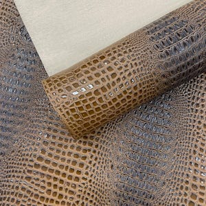 May include: A close-up of a roll of brown and gray faux crocodile leather. The leather has a glossy finish and a textured, scale-like pattern. A piece of off-white fabric is visible in the background.
