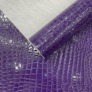 May include: A roll of purple faux leather with a glossy, crocodile-skin texture. The material is partially unrolled, revealing a grey, woven backing. The surface reflects light, highlighting the embossed pattern.