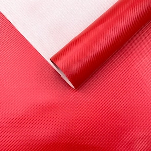 May include: A roll of vibrant red carbon fibre vinyl wrap material. The material features a textured, diagonal weave pattern. A section of the material is unrolled, revealing a white backing. The roll is positioned diagonally across the frame.