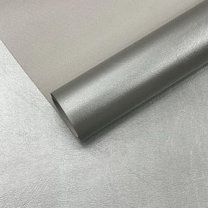 Può includere: Un rotolo di materiale in similpelle grigio argento. Il materiale ha una superficie strutturata ed è parzialmente arrotolato, rivelando il bordo. Lo sfondo mostra un foglio piatto dello stesso materiale.