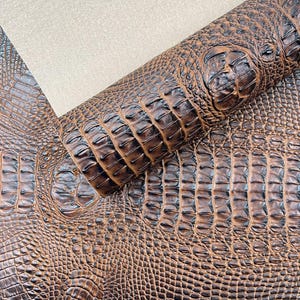 Peut inclure: Gros plan sur du cuir marron texturé façon crocodile. Le cuir est d'un brun foncé riche avec une finition brillante, mettant en valeur le motif complexe d'écailles et d'arêtes. Une section roulée du cuir est visible, révélant la texture et la profondeur du matériau.