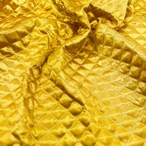 Op de afbeelding: Close-up van een goudgeel gewatteerde stof. De stof heeft een ruitpatroon dat is gemaakt door gestikte lijnen. Het materiaal heeft een glanzende, satijnachtige uitstraling, met plooien en schaduwen die diepte en textuur toevoegen.