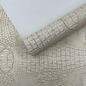 Puede incluir: Cuero texturizado blanco roto, imitando la piel de cocodrilo, parcialmente enrollado, con un patrón detallado de escamas y crestas. El material tiene un brillo sutil y la textura es visible. Fondo blanco liso.
