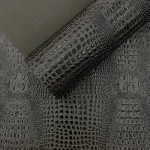 Może przedstawiać: Zbliżenie czarnej skóry ekologicznej imitującej krokodyla. Materiał jest błyszczący i ma powtarzający się wzór łusek. Widoczny jest zwinięty fragment skóry, ukazujący fakturę i połysk. Całość daje luksusową, teksturowaną powierzchnię.