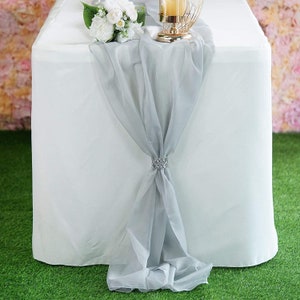 May include: A white tablecloth with a silver chiffon table runner tied with a silver rhinestone embellishment. The runner is draped over the table and hangs down the sides.
