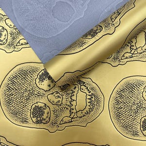 May include: Gold-colored wrapping paper with a black skull pattern. A rolled section of the paper reveals the pattern, while a flat section shows the repeating design. A light blue fabric with a skull pattern is visible in the background.