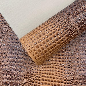 May include: A roll of brown faux leather with a crocodile skin texture. The material has a glossy finish and varying shades of brown, creating a realistic appearance. A section of beige fabric is visible in the background.