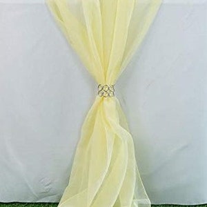 May include: A pale yellow sheer table runner with a decorative silver ring tied around it. The runner is draped over a white tablecloth.