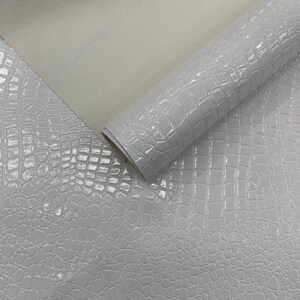 Pode incluir: Um rolo de couro sintético branco com uma textura brilhante de pele de crocodilo. O material está parcialmente desenrolado, revelando um verso liso e fosco. A superfície reflete a luz, destacando o padrão em relevo.