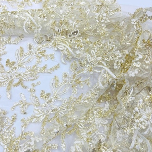 May include: Close-up of a delicate, sheer fabric with intricate floral embroidery. The fabric is a soft ivory color, embellished with gold sequins and thread, creating a shimmering effect. The design features detailed floral patterns.