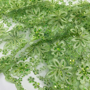 Op de afbeelding: Close-up van een lichtgroene bloemenkanten stof. De stof heeft geborduurde bloemen en bladeren met glinsterende pailletten. Het kant is gelaagd, waardoor een textuur en dimensionaal effect ontstaat. Het ontwerp is ingewikkeld en gedetailleerd, geschikt voor kleding en knutselprojecten.