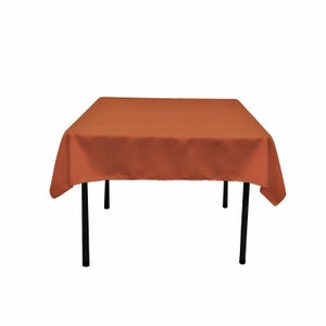 May include: A square, rust-colored tablecloth draped over a black metal table. The tablecloth is made of a smooth fabric and covers the entire table surface. The table has four black legs.
