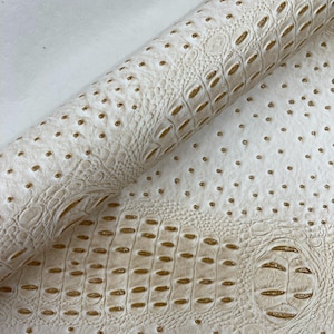 May include: Cream-colored faux leather material with a crocodile and ostrich skin pattern. The material is rolled up, revealing the textured surface and the detailed design. The color is a soft off-white with subtle golden accents.