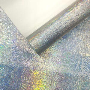 Puede incluir: Primer plano de tela plateada iridiscente con textura arrugada. La tela está parcialmente enrollada, revelando un reverso blanco. La superficie refleja un espectro de colores, creando un efecto brillante. Ideal para proyectos de manualidades y diseño.