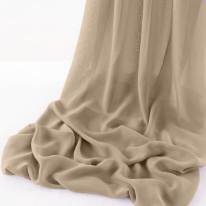 May include: A flowing, beige fabric with a soft, translucent appearance. The material is draped and folded, creating gentle waves and shadows. The fabric appears lightweight and airy, suggesting it could be used for curtains or apparel.