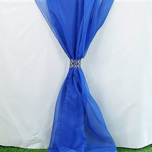 Pode incluir: Um corredor de mesa transparente azul real com um laço de cristal de strass. O corredor de mesa está drapeado sobre uma toalha de mesa branca e está sobre uma superfície de grama verde.