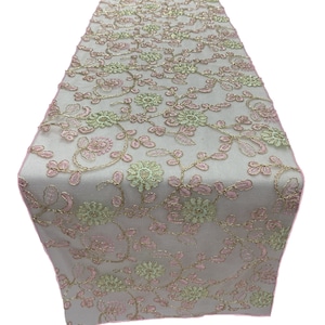 May include: A delicate white table runner with a floral design featuring pink and green embroidered flowers and vines. The runner has a subtle shimmer and a pink border.