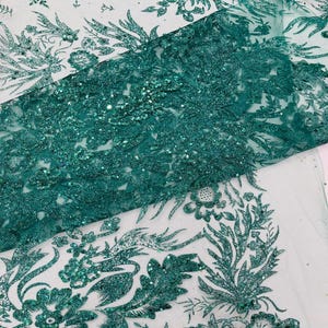 Op de afbeelding: Close-up van smaragdgroene kanten stof met bloemen- en bladerpatronen. De stof is versierd met pailletten, wat een glinsterend effect geeft. Het ontwerp heeft gedetailleerd borduurwerk en een delicate, open weefsel, geschikt voor kleding en knutselprojecten.