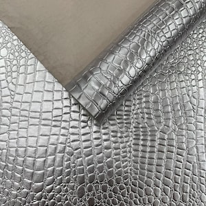 May include: Silver faux leather material with a crocodile skin texture. The material is rolled up and partially unrolled, revealing a smooth, light-colored backing. The surface has a metallic sheen, reflecting light.