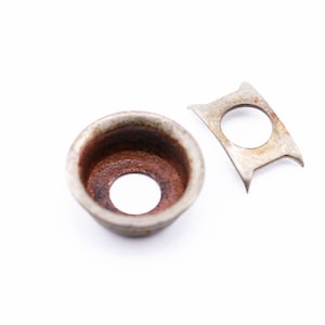 May include: A close-up shot of two aged metal components. One is a circular, cup-shaped piece with a central hole and a rusty interior. The other is a small, irregularly shaped piece with a circular hole. Both pieces have a worn, aged appearance.
