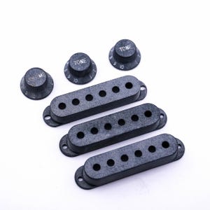 Könnte beinhalten: Drei schwarze Gitarren-Tonabnehmerabdeckungen und drei schwarze Knöpfe mit weißer Beschriftung. Die Knöpfe sind mit "TONE" und "VOL. M" beschriftet.