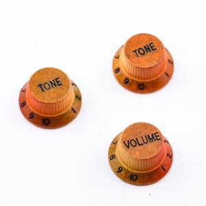 Könnte beinhalten: Drei Gitarrenknöpfe in einem verblassten Orange- und Braunton. Die Knöpfe sind mit "TONE" und "VOLUME" beschriftet und haben die Zahlen 1-10 am Rand.