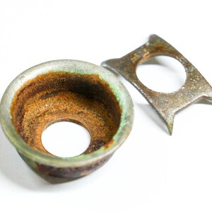 May include: Close-up of two antique metal objects with a weathered appearance. One is a bowl-shaped piece with a central hole and a green-tinged rim. The other is a flat, angular piece with a circular opening and pointed ends.