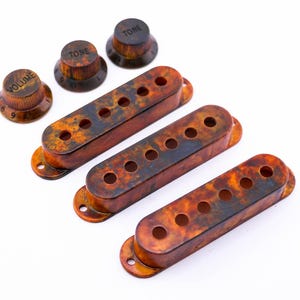 Könnte beinhalten: Vier Gitarren-Pickup-Abdeckungen in einem gefleckten Orange- und Braunton. Drei Knöpfe mit den Worten "Volume", "Tone" und "Tone" sind ebenfalls enthalten.