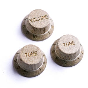 Może przedstawiać: Trzy beżowe pokrętła do gitary z nadrukowanymi słowami "VOLUME" i "TONE". Pokrętła mają liczby od 1 do 10 wokół podstawy. Pokrętła są wykonane z teksturowanego materiału i są umieszczone na białym tle.