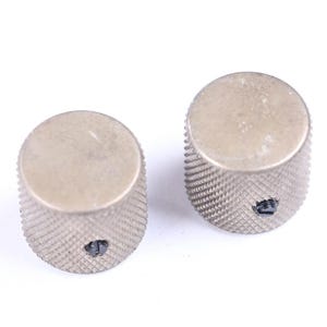 May include: Two silver-colored control knobs with a textured, ridged surface. The knobs have a circular top and a small set screw. These knobs are likely for musical instruments or electronic devices.