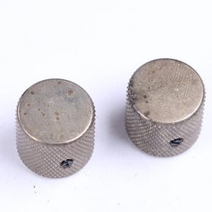 May include: Two silver-colored control knobs with a textured, ridged design on the sides. The tops of the knobs are flat and show signs of wear. Each knob has a small, dark hole.