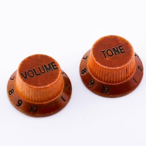 Könnte beinhalten: Zwei braune Gitarrenknöpfe mit schwarzen Zahlen und Beschriftung. Der Knopf links sagt "VOLUME" und der Knopf rechts sagt "TONE".