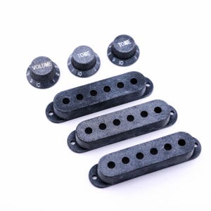 Könnte beinhalten: Drei schwarze Kunststoff-Tonabnehmerabdeckungen und drei schwarze Knöpfe mit weißer Beschriftung. Die Knöpfe zeigen "Volume", "Tone" und "Tone".