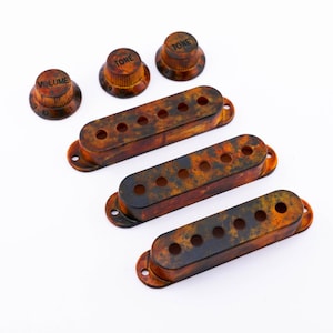 Może przedstawiać: Zestaw trzech osłon pickupa do gitary w kolorze żółwim z sześcioma otworami każda i trzema pokrętłami oznaczonymi "Volume" i "Tone".