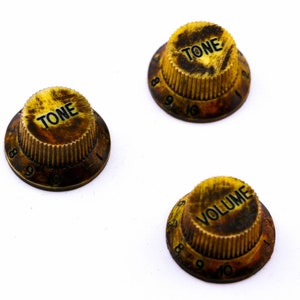 Könnte beinhalten: Drei goldfarbene Gitarrenknöpfe mit schwarzer Beschriftung. Die Knöpfe sind mit "TONE" und "VOLUME" beschriftet.