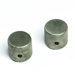 May include: Two silver-colored, cylindrical knobs with a textured, diamond-patterned grip. Each knob has a flat top and a small, dark screw visible on the side. The knobs are likely for a musical instrument or electronic device.
