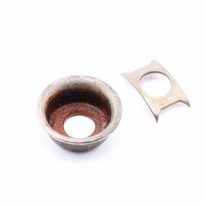 May include: A close-up of two metal objects on a white background. One is a small, round, silver-colored cup with a rusty brown interior. The other is a small, silver-colored, curved piece with a circular hole in the center.