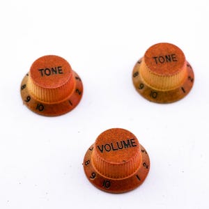 Könnte beinhalten: Drei Gitarrenknöpfe in orange und brauner Farbe. Die Knöpfe sind mit "TONE" und "VOLUME" beschriftet und haben die Zahlen 1 bis 10 am Rand.