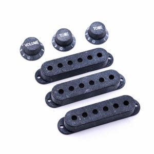 Könnte beinhalten: Ein Satz schwarzer Gitarrenteile, darunter drei Tonabnehmer mit jeweils sechs Löchern und drei Bedienknöpfe. Die Knöpfe sind mit "VOLUME" und "TONE" sowie den Zahlen 9 und 10 beschriftet. Die Teile liegen auf weißem Hintergrund.